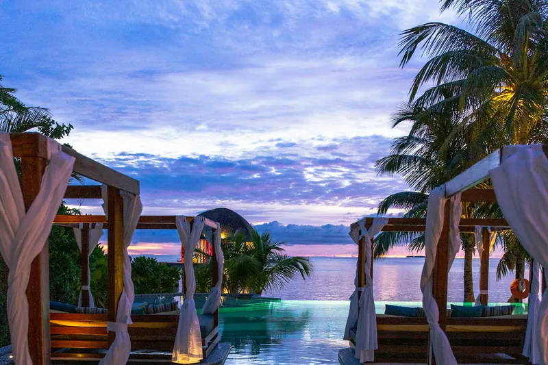 Timber shade structures with draped fabric beside a pool at dusk.