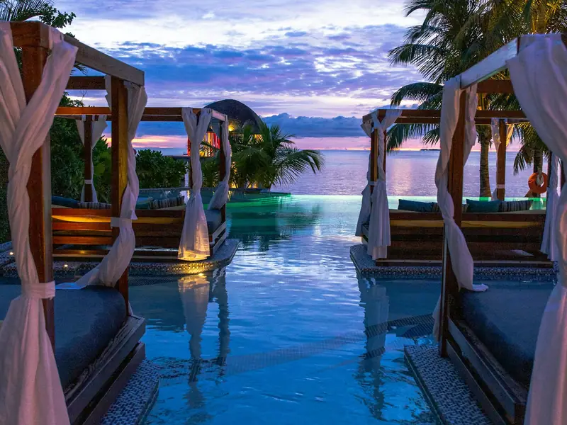 Timber shade structures with draped fabric standing beside a lit pool at dusk.