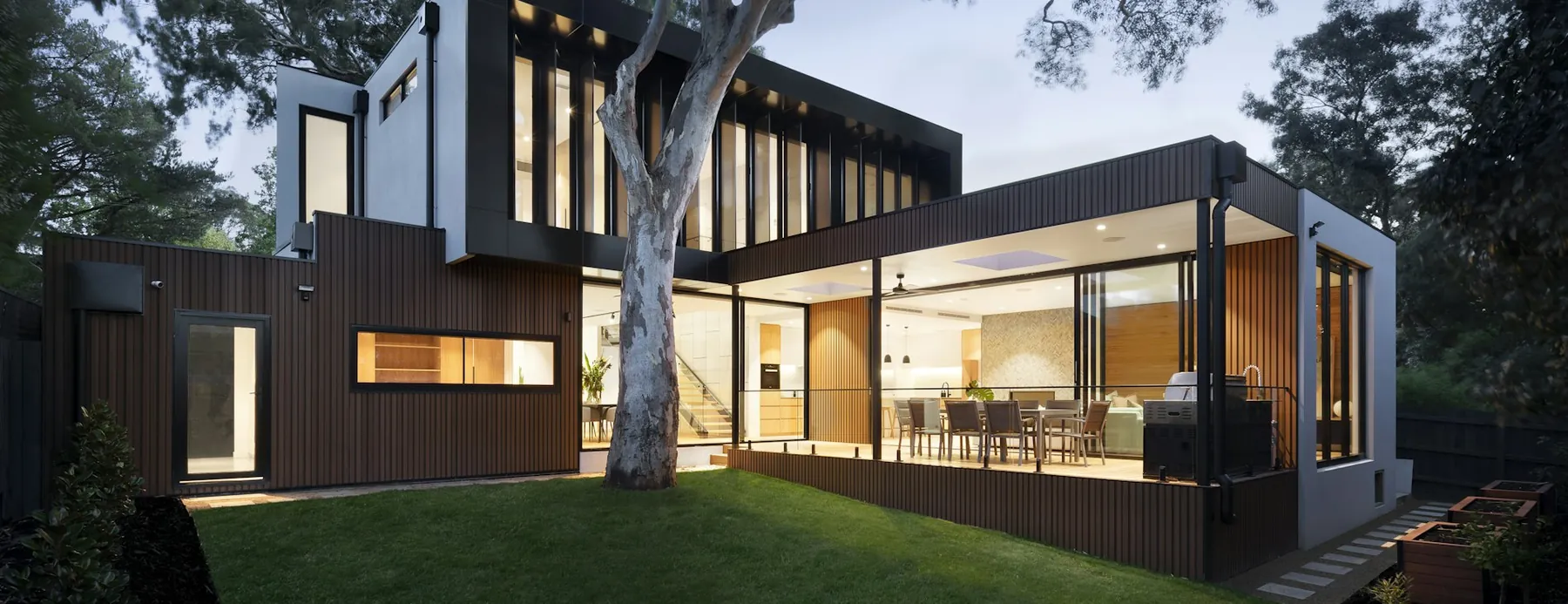 Covered terrace with a dining setting opening through full-height glazing onto a level lawn at dusk.